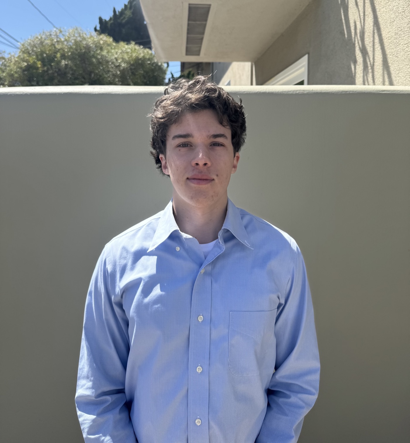 Zo Keck-Choeff, photographed outdoors in bright sun against a pale stucco wall, wearing a light blue button-down shirt.